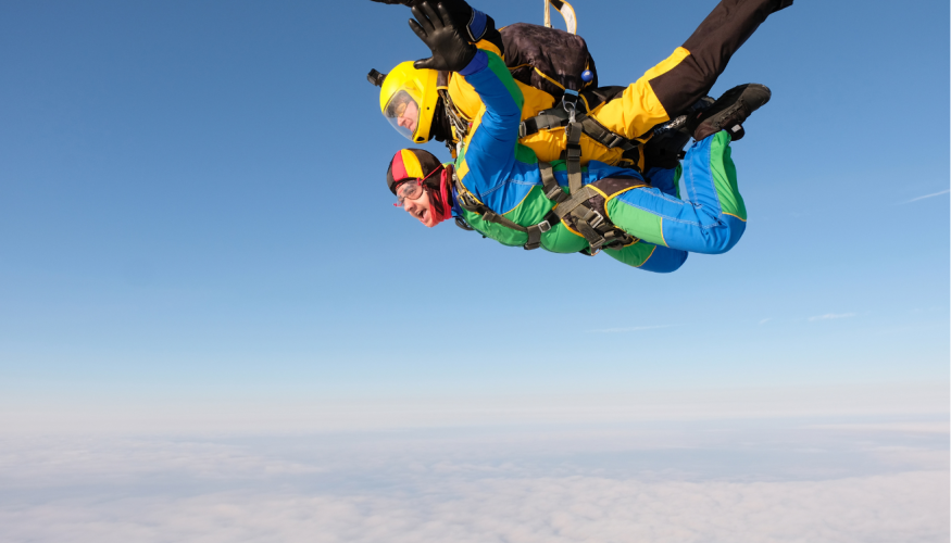 Kaksi ihmistä tandemhyppäämässä kädet ja jalat levällään värikkäissä haalareissa